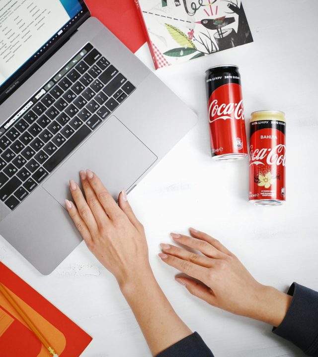 Top view of a workspace with hands on a laptop and Coca-Cola cans nearby.
